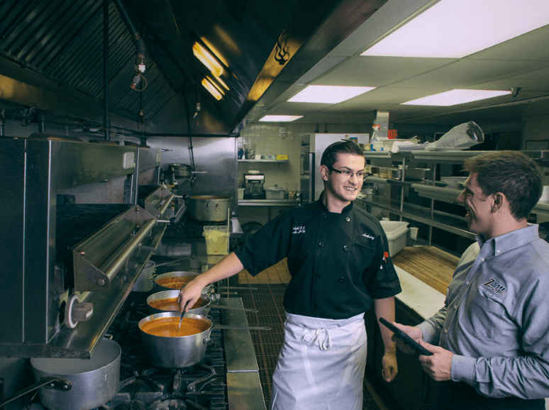 A Zoom Drain technician talks to a cook stirring a pot in a commercial kitchen