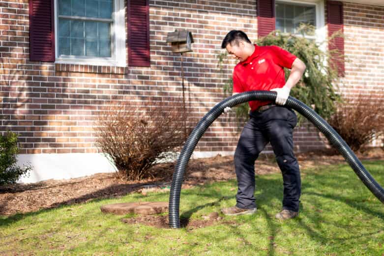 A Zoom Drain technician performs trenchless sewer repair by guiding a flexible black tube into a small hole in the ground in front of a house